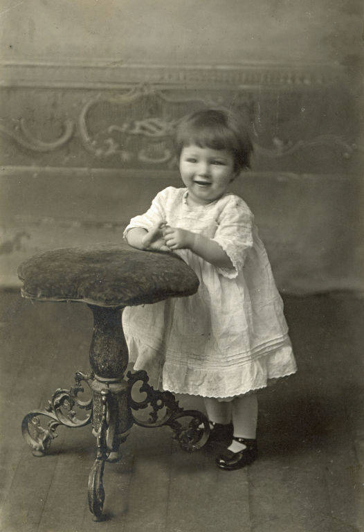 A young child in a white dress and dark shoes stands next to a cushioned stool with ornate legs. The child is smiling and has their hand resting on the stool. The background features decorative patterns.