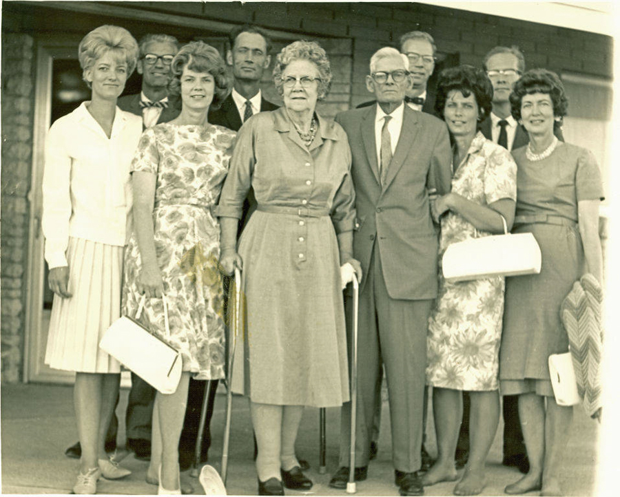 A group of nine people is standing together outside a building. In the front, an elderly couple is centered; the woman holds a cane. The group includes men in suits and women in dresses, some holding handbags. The women have various hairstyles and some wear patterned dresses. The setting appears formal.