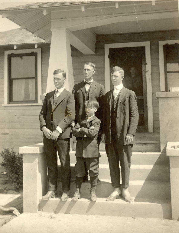 Four young males in formal suits stand on the steps of a house. A small boy is in front of the three older individuals. The house has a porch with a large column and a person is partially visible behind a screen door in the background.