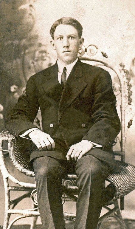 A man wearing a dark suit and tie sits upright in an ornate wicker chair. The background features a subtle decorative pattern. His hands rest on his lap.