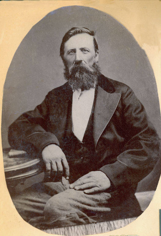 A man with a beard is seated, wearing a suit with a vest and resting his arm on a table.