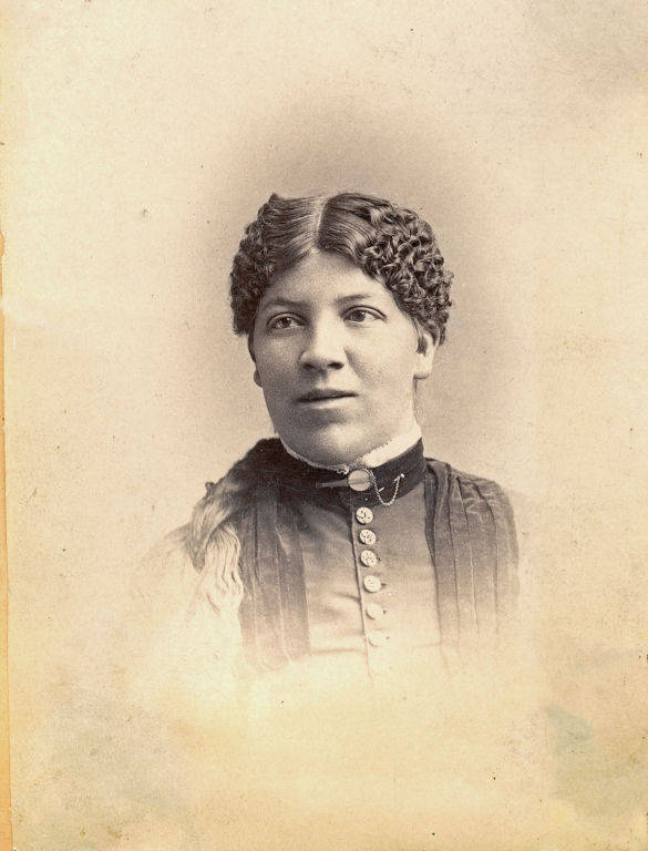 A woman with tightly curled hair gazes to her left. She is wearing a dark dress with vertical pleats, a row of buttons down the front, and a high collar. Her expression is neutral, and her hair is parted in the middle.