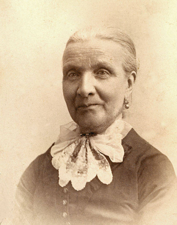 An older woman with her hair tied back, wearing a dress with buttons and a lace collar. She also has earrings and is looking slightly to the side.