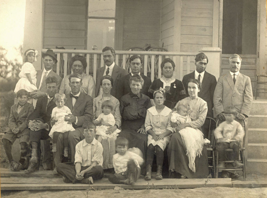 A large group of mostly adults and a few children are seated and standing, posing together in front of a house with stairs and a porch. The group includes a mix of men, women, and children, with some children sitting on laps. The individuals are dressed in formal attire typical of an earlier time period. There is no visible text in the image.