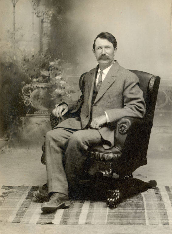 A man with a mustache sitting in an ornate armchair, wearing a suit and tie. The chair is on a patterned rug, and the background shows a decorative setting with plants and an arched window.