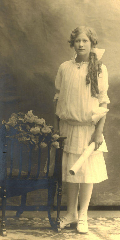 A young girl with long hair standing, wearing a light-colored dress and holding a rolled-up document in her right hand. She is standing next to a chair with a floral arrangement on it.