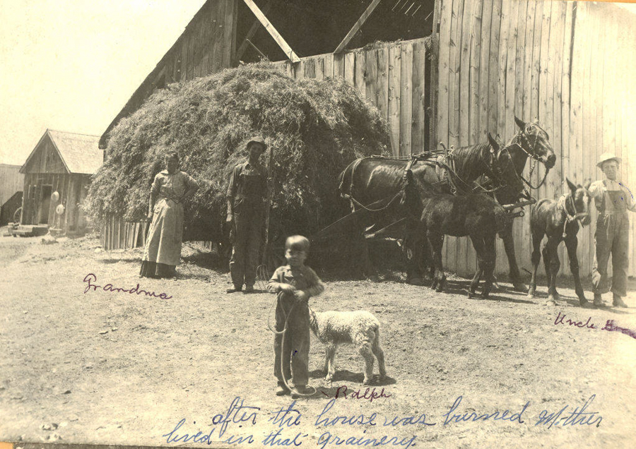 A young child stands in front of a small animal, possibly a sheep, in a farmyard. A woman labeled "Grandma" stands near a large pile of hay. Two horses are hitched to a wagon, with another child, labeled "Uncle Earl," standing beside them. A man in work clothes stands next to the woman. Buildings are visible in the background. Handwritten text reads, "Ralph" near the child, and "after the house was burned Mother lived in that granary."