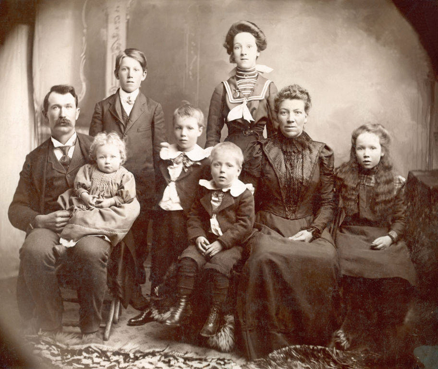 A group of eight people, consisting of two adults and six children, is arranged in a studio setting. The adults, a man on the left holding a child and a woman on the right, are seated. The children are positioned both seated and standing. The children wear outfits with large collars and bows, with two boys in the front and two girls on either side. The backdrop suggests an interior, and the floor is covered with a patterned rug. The group faces forward with neutral expressions.