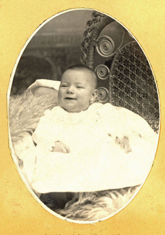 A baby lying on a textured fabric in a wicker chair with a decorative backrest. The baby is wearing a white gown and appears to be smiling or content.