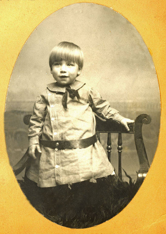 A young child standing on a chair, wearing a buttoned-up dress with a collar and a bow, and a wide belt. The child has short hair and is holding onto the chair's armrest.