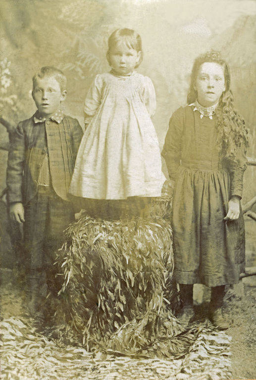 Three children are posed together. A young girl in a dress stands on a chair covered with foliage in the center. To her right, a girl with long hair and a patterned dress stands. To her left, a boy in a suit with a bow tie stands. The backdrop includes foliage and branches.