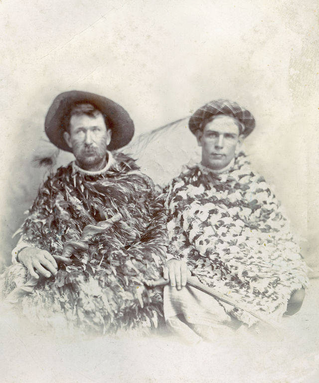Two men sitting side by side, each wearing a cloak made of feathers. The man on the left has a beard and is wearing a wide-brimmed hat. The man on the right has short hair and is wearing a patterned cap. Both are looking towards the viewer with a neutral expression.