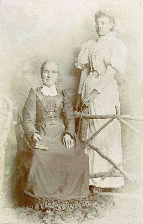 An older woman is seated with a book on her lap, wearing a long dress with a high collar and buttons down the front. A younger woman stands beside her, dressed in a similar long, elegant dress with puffed sleeves and a narrow waist. They are positioned near a rustic wooden fence.