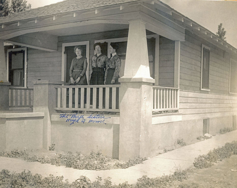 Three women standing on the porch of a wooden house with a low railing and pillar. The ground is covered with grass and a cement pathway leads to the porch. Handwritten text says "The Three Sisters High & Home."