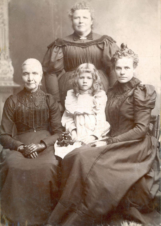 Four women and a girl are posed together. The woman on the left is seated, wearing gloves and a dark dress with beaded details. The girl in front is sitting with loose hair and a white dress, holding flowers. The woman on the right is also seated and dressed in a dark, formal gown. Standing behind them is another woman wearing a dark dress with puffed sleeves. All appear serious or solemn in expression.