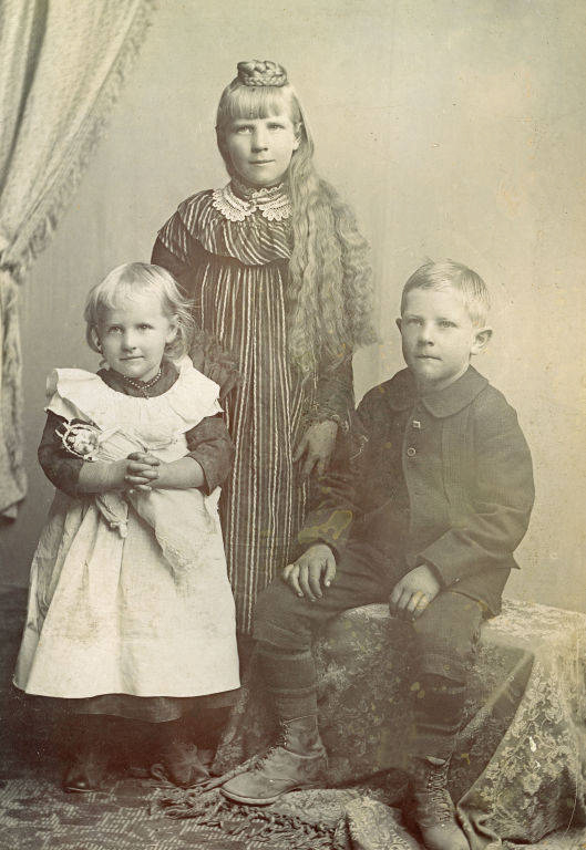A young girl with long hair stands behind two younger children. The girl is wearing a striped dress with a lace collar. One child is a girl holding a doll, wearing a white pinafore over a dark dress. The other child is a boy seated on a patterned chair, wearing a dark suit with buttons. A curtain is draped in the background.