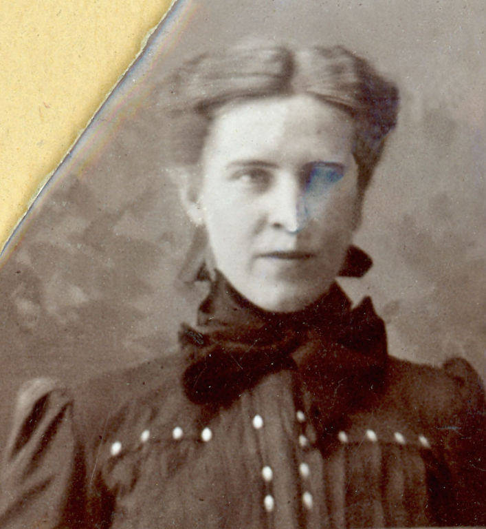 A woman with her hair neatly styled is wearing a high-collared dress with a row of buttons down the front. The background has a subtle pattern, and there is a small discolored area over her right eye. The top left corner of the image is irregular, possibly due to an old paper or mounting.