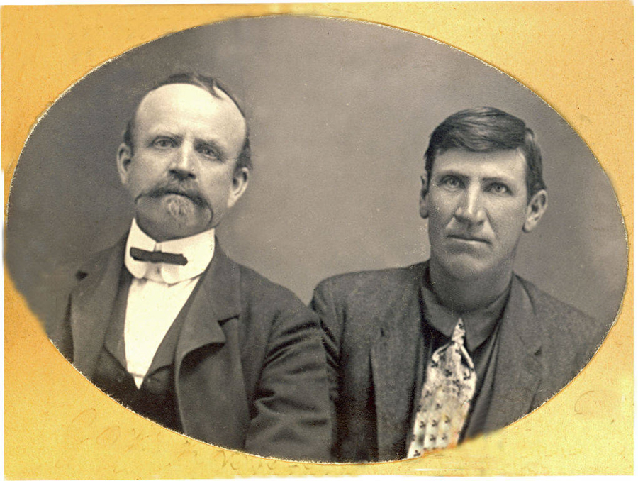 Two men seated side by side, both wearing suits. The man on the left has a mustache and bow tie, while the man on the right has a patterned tie. The background is plain.
