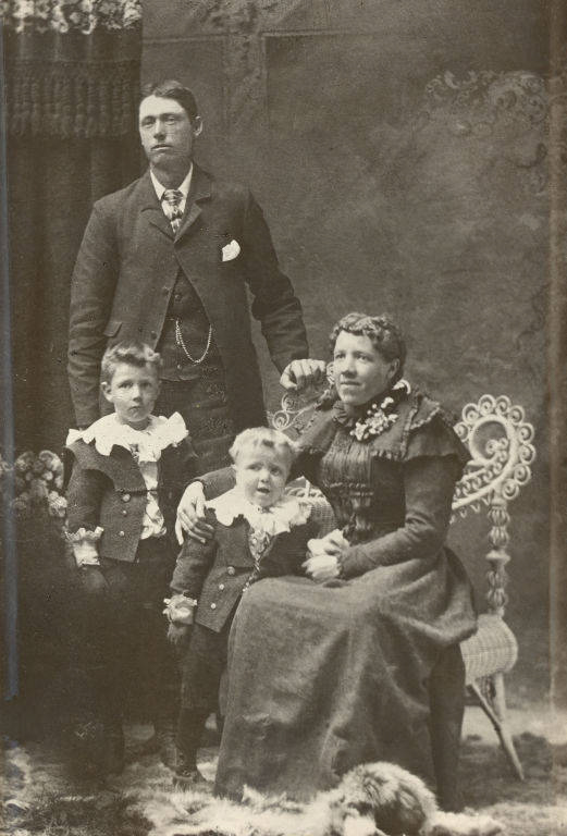 A man stands beside a seated woman, both dressed in formal attire. The woman has her arm around a young child, who stands beside her. Another young child stands next to the man. Both children wear matching outfits with white collars. The group is posed in front of a decorative backdrop with a patterned chair. A small dog lies on the ground in front of them.