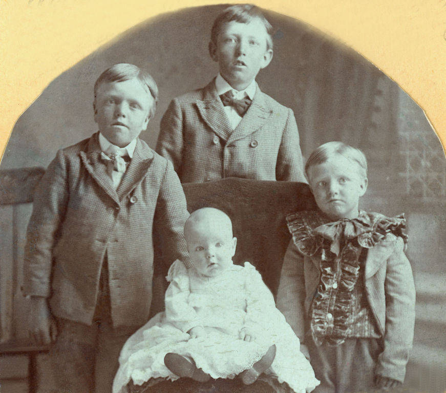 Four young children are posed together. Three of them stand, wearing suits with bow ties or cravats. A baby sits in front wearing a white gown. The children are arranged in a formal setting.