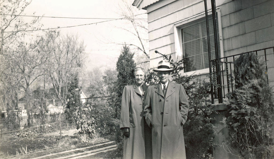 A man and a woman standing side by side in front of a house. Both are wearing long coats, and the man is wearing a hat. There are shrubs and trees around them, with a garden and fence visible in the background.