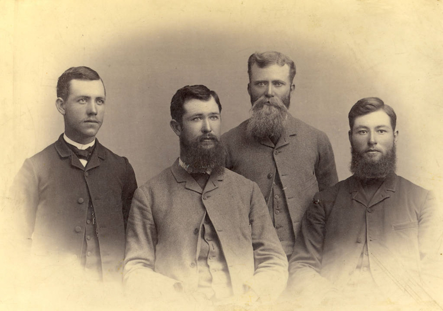Four men with serious expressions are posed together. Three of them have full beards, while one is clean-shaven. They are dressed in formal jackets and buttoned shirts, with two standing at the back and two sitting in front.
