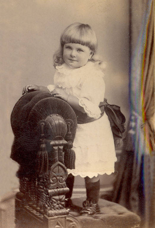 A young child with light hair stands on a cushioned chair adorned with ornate carvings and tassels. They are dressed in a white outfit with lace details and a dark sash. The child is gazing slightly towards the side.