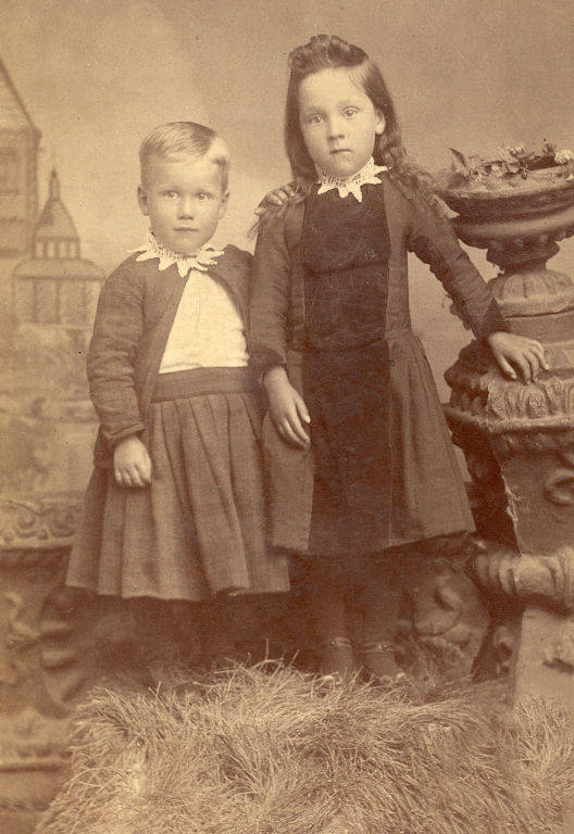 Two young children stand side by side, both wearing clothing with lace collars. The child on the left wears a lighter outfit and has short hair, while the child on the right has longer hair and wears a darker dress. They stand in front of a backdrop with architectural elements and next to a decorative stone structure with plants. The ground is covered with hay or straw.