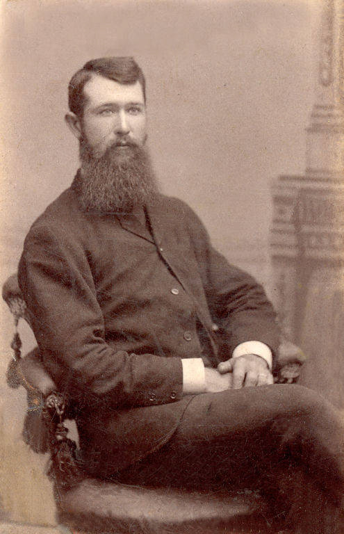 A man with a long beard is seated on an ornate chair. He is wearing a dark suit with a high collar and has his hands resting on his lap. There is a partially visible decorative column in the background.