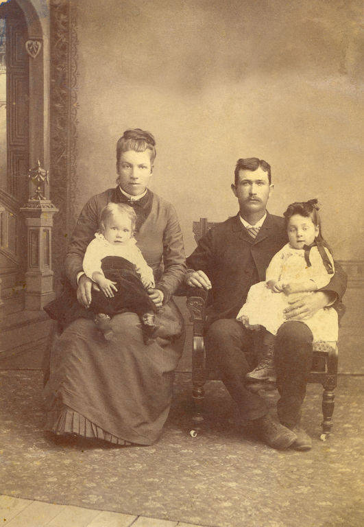 A man and woman are seated in chairs next to each other. The woman is holding a child on her lap, and the man is holding another child on his lap. The setting is indoors, with a patterned carpet and a decorative archway in the background. The woman and children are wearing dresses, and the man is dressed in a suit.