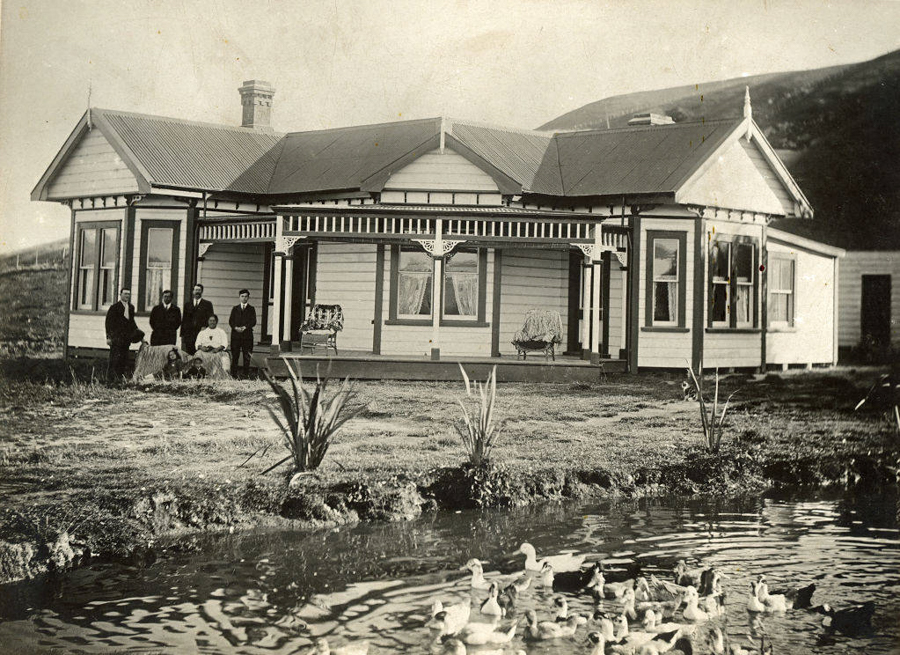 A group of people stands in front of a wooden house with a veranda and decorative trim. There are several adults and a couple of children, some sitting and some standing. In the foreground, there is a pond with numerous ducks swimming. The landscape around the house is open and slightly hilly.