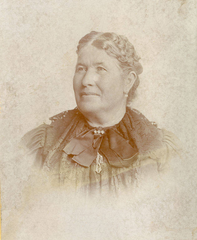 An older woman with coiffed hair looks slightly upwards. She is wearing a dark, textured dress with a large bow at the collar and earrings. The background is plain.