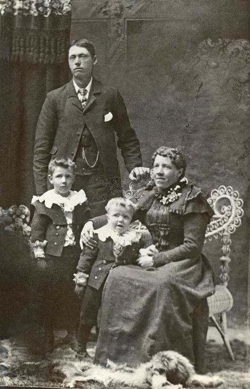 A man stands behind a seated woman who is holding the hands of two young children. The man is dressed in a suit and the woman is wearing a long dress with lace detailing. The children are also in suits with large collars. The woman is seated on an ornate chair, and there is a small dog or animal lying on the floor in front of them. The background features a decorative draped curtain.