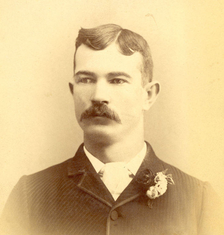 A man with a mustache wearing a suit and tie, with a floral boutonniere on his lapel. His hair is neatly parted and combed to the side. He has a serious expression and is facing forward.