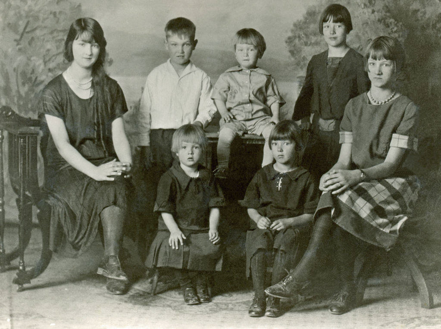A group of seven children sit and stand in a formal arrangement with a backdrop resembling a garden landscape. The children are dressed in early 20th-century attire. Two are seated on either side, while five are positioned in the middle, with two sitting on a bench. The expressions are neutral.
