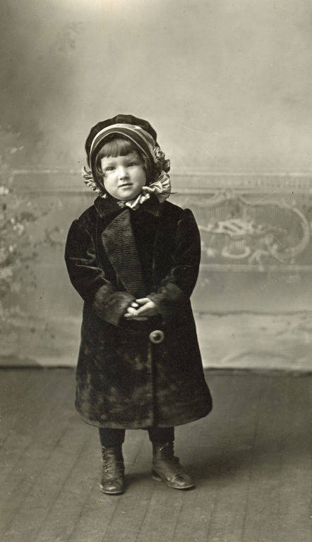A young child standing on a wooden floor wearing a dark, heavy coat, buttoned up, and a bonnet with ruffled edges. The child's hands are clasped in front, and they appear to be looking forward. The background has a decorative design resembling a wall or screen.