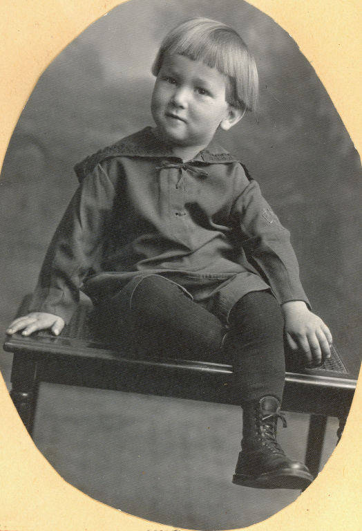 A young child sitting on a small table, wearing a tunic with a collar, leggings, and lace-up boots. The child has short, straight hair and is looking slightly off to the side.