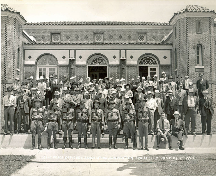 A large group of people, including men and women, stand in front of a brick building with arched windows and ornate details. Many of the men wear suits and hats, while several individuals in the front row are in uniform. The text at the bottom reads, "Idaho Peace Officers Association Convention Pocatello June 24-25 1940."
