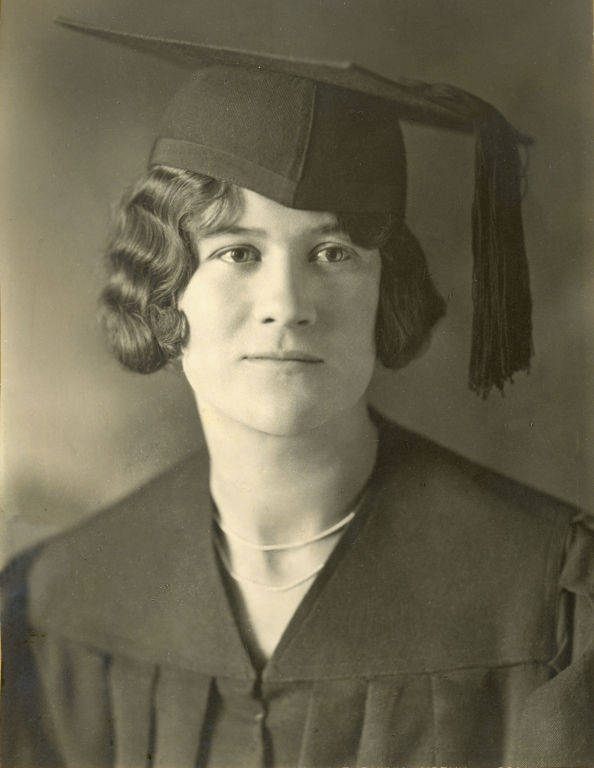 A woman wearing a graduation cap and gown with short, wavy hair. She is looking directly at the viewer with a neutral expression.