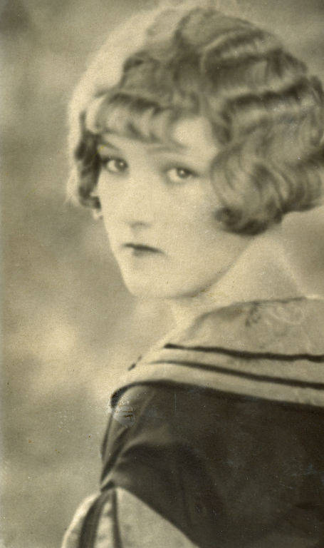 A person with short, wavy hair is looking over their shoulder. They are wearing a garment with a visible collar and decorative trim. The expression is neutral and the background is plain.