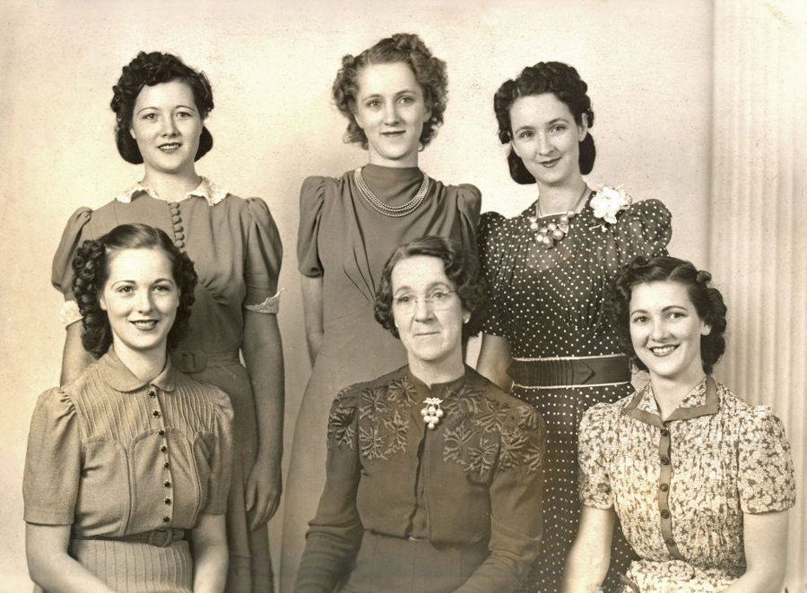 Six women are posed in two rows, with three standing at the back and three seated in front. They are wearing vintage dresses with distinct patterns and styles. A woman in the center of the front row is wearing glasses and a dress with floral embroidery. The others have various hairstyles typical of an earlier era. There is no text visible.