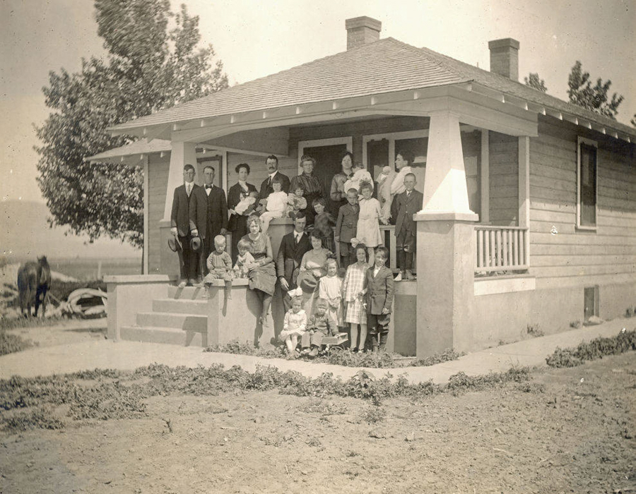 A large group of people is gathered on the porch of a house. The group includes men, women, and children of various ages, with some standing and others sitting on the steps. There is a tree beside the house, and a horse is visible in the background. The house has a gabled roof and wooden siding.