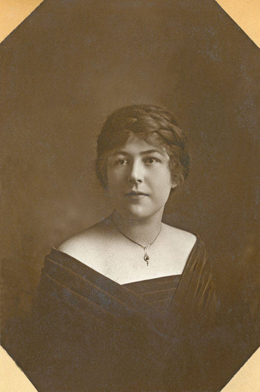 A woman with short, styled hair is wearing an off-the-shoulder dress and a necklace with a small pendant. She is looking slightly to the side. The background is plain and dark.