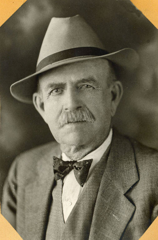 An older man wearing a suit, bow tie, and a wide-brimmed hat. He has a mustache and is looking directly at the viewer with a neutral expression.