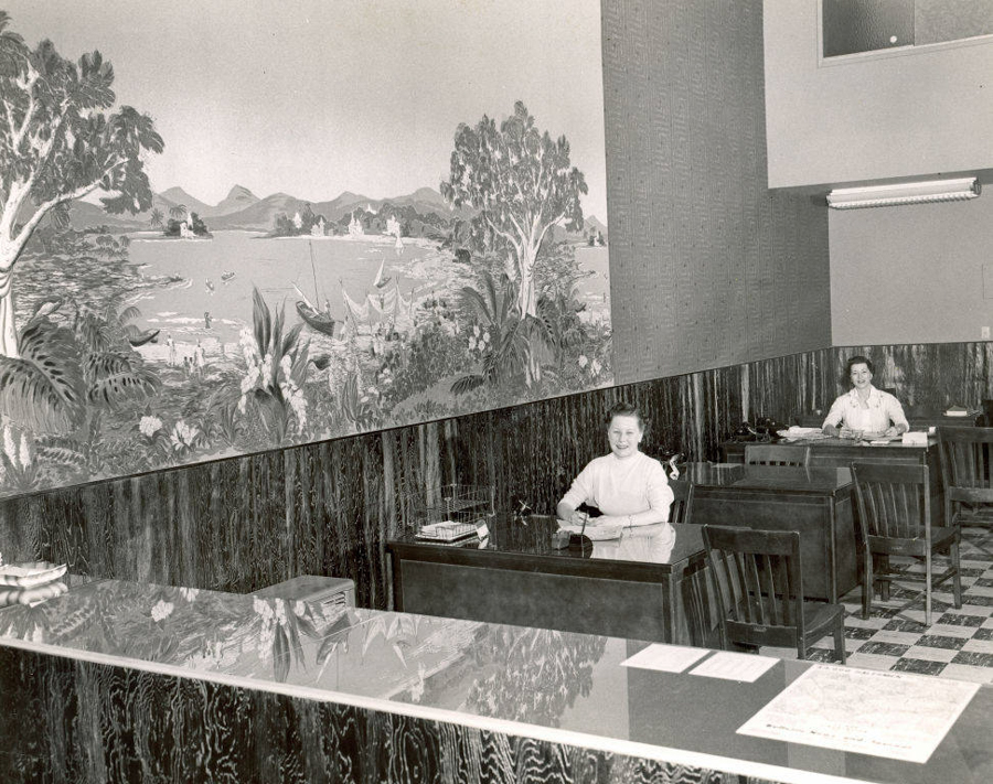 Two women are seated at separate desks in a room. The woman closer to the foreground is smiling, while the other is sitting at a desk further back, partially covered by papers. The desk in the foreground has a phone on it. A mural depicting a landscape with trees, mountains, and a body of water with boats covers the wall behind them. The floor is checkered, and there is a countertop in the foreground with papers on it.