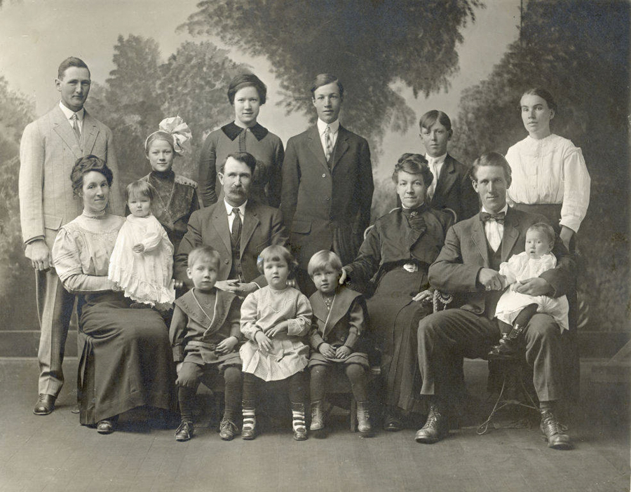 A group of fourteen people is posed in three rows. In the back row, there are four men and two women standing, all dressed in formal attire. In the middle row, three men and one woman are seated, with the woman holding a baby. In the front row, four young children are seated, with two girls and two boys. The scene appears to be a formal family portrait against a painted backdrop.