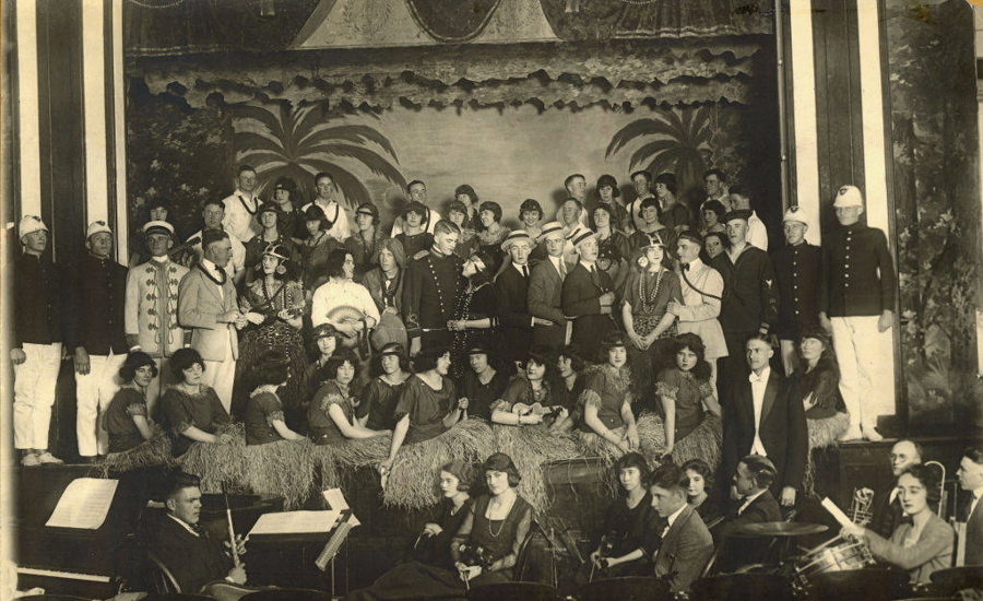 A large group of men and women are gathered on a stage. The men are dressed in various uniforms and suits, while the women wear costumes made from grass skirts and other tropical-themed outfits. Some individuals are holding musical instruments, and an orchestra is visible in the foreground. The backdrop features a tropical scene with palm trees.