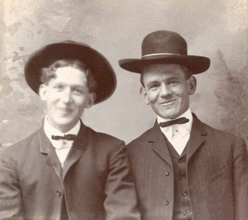 Two men are standing side by side, both wearing dark suits, white shirts, bow ties, and hats. They are smiling and appear to be posing for the camera with a plain background behind them.