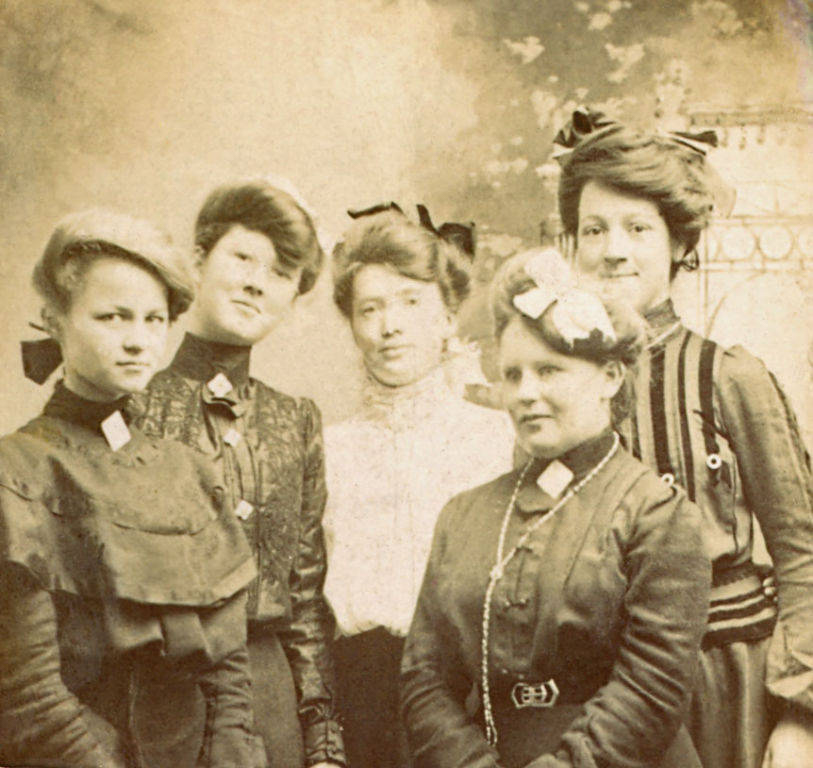 Five women are standing closely together. They are wearing high-collared dresses with decorative patterns. Their hairstyles are styled up and adorned with bows and ribbons. The background is decorated with a painted or textured wall.