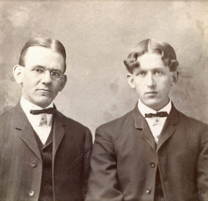 Two men seated and facing forward. The man on the left wears glasses and a suit with a bow tie. The man on the right also wears a suit with a bow tie. Both have neatly styled hair and a serious expression.